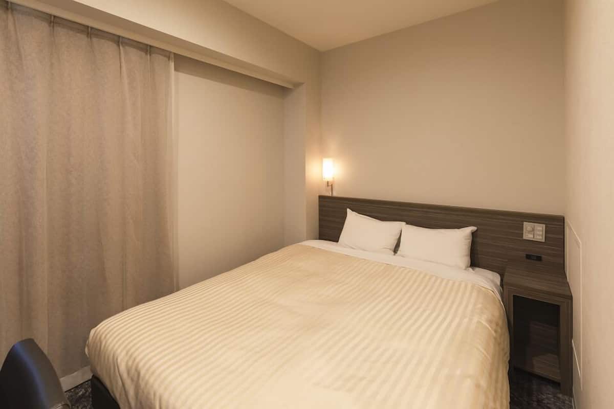 Quarto de hotel com cama, mesa de cabeceira, luminária e janela com cortinas.