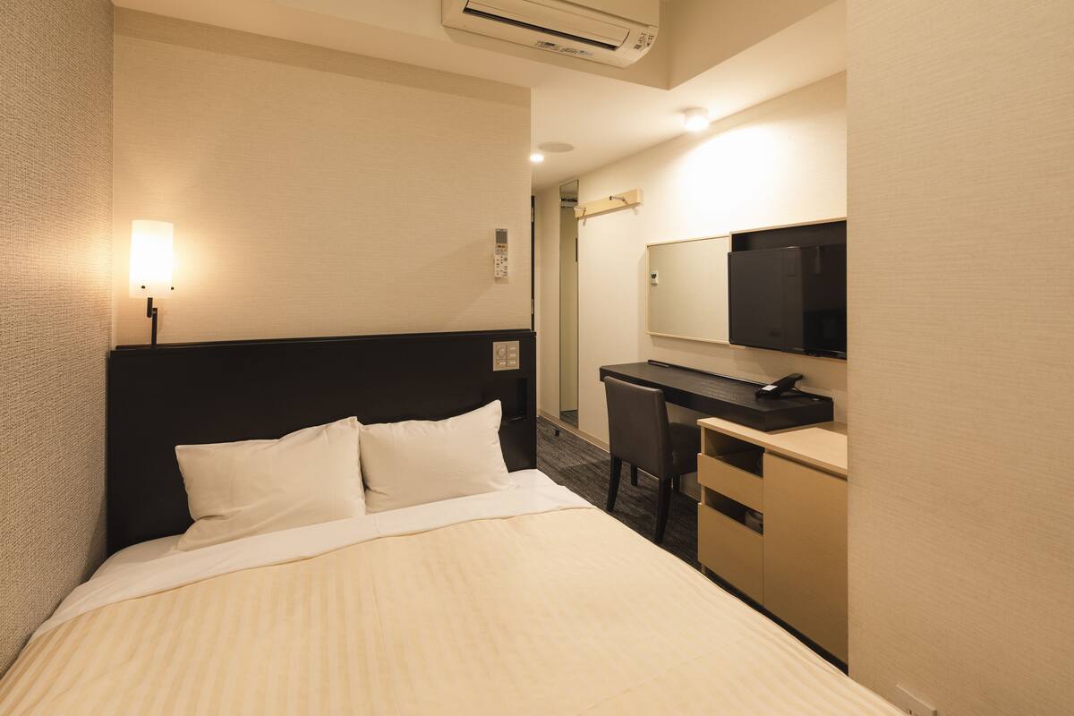 A hotel room with a bed, a desk, a chair, a mirror, and a wall-mounted TV.
