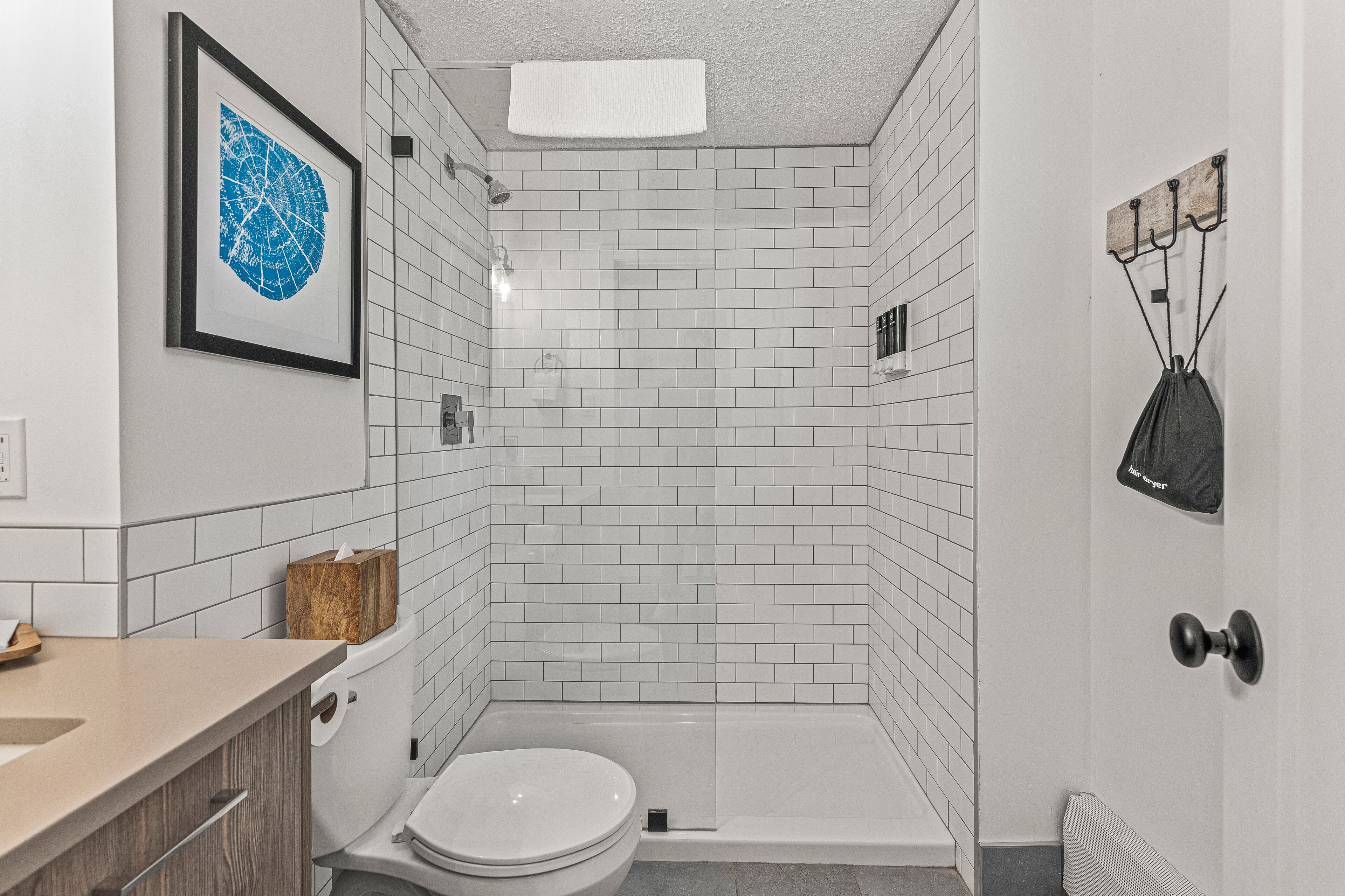 Une salle de bain moderne avec des carreaux de céramique blancs rectangulaires, une œuvre d  ’art encadrée, une boîte à mouchoirs en bois et un espace douche.