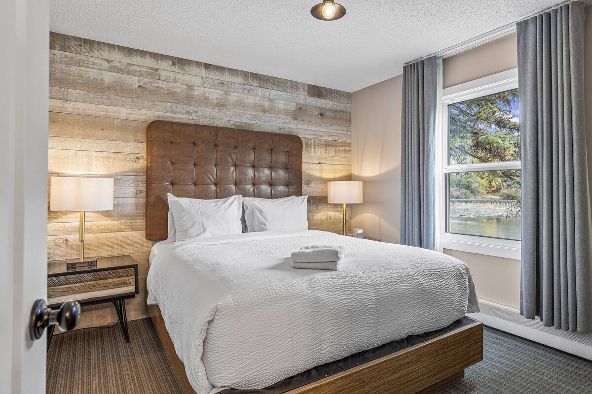 A hotel room with a wooden headboard, a large window with curtains, a bed with white linens, and two bedside lamps.