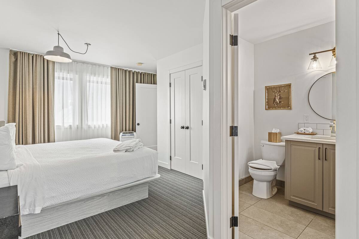A hotel room with a bed, a bedside table, a window with curtains, a bathroom with a toilet, sink, and mirror.