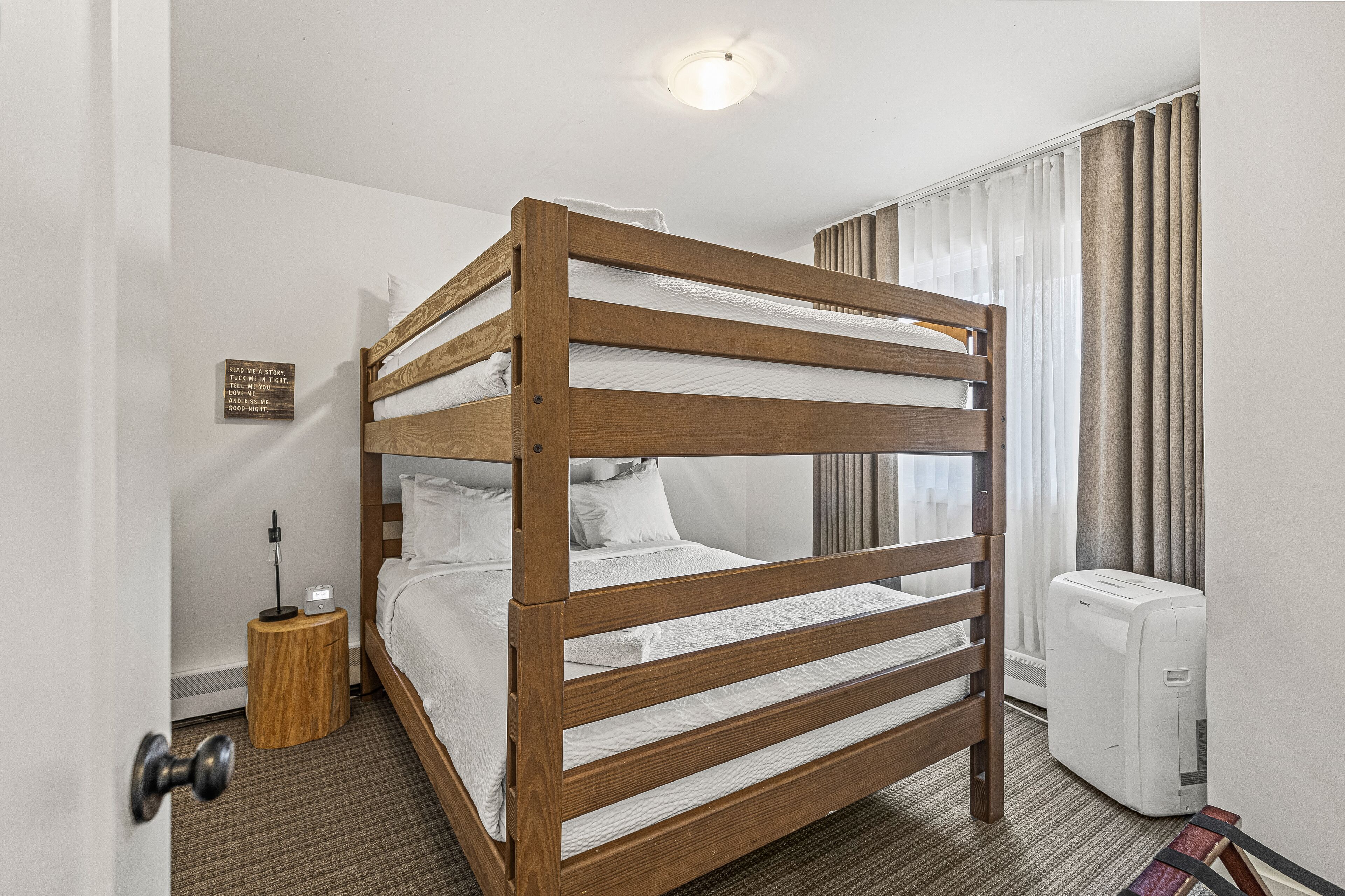 A bunk bed in a hotel room with a bedside table, a lamp, and an air conditioning unit.