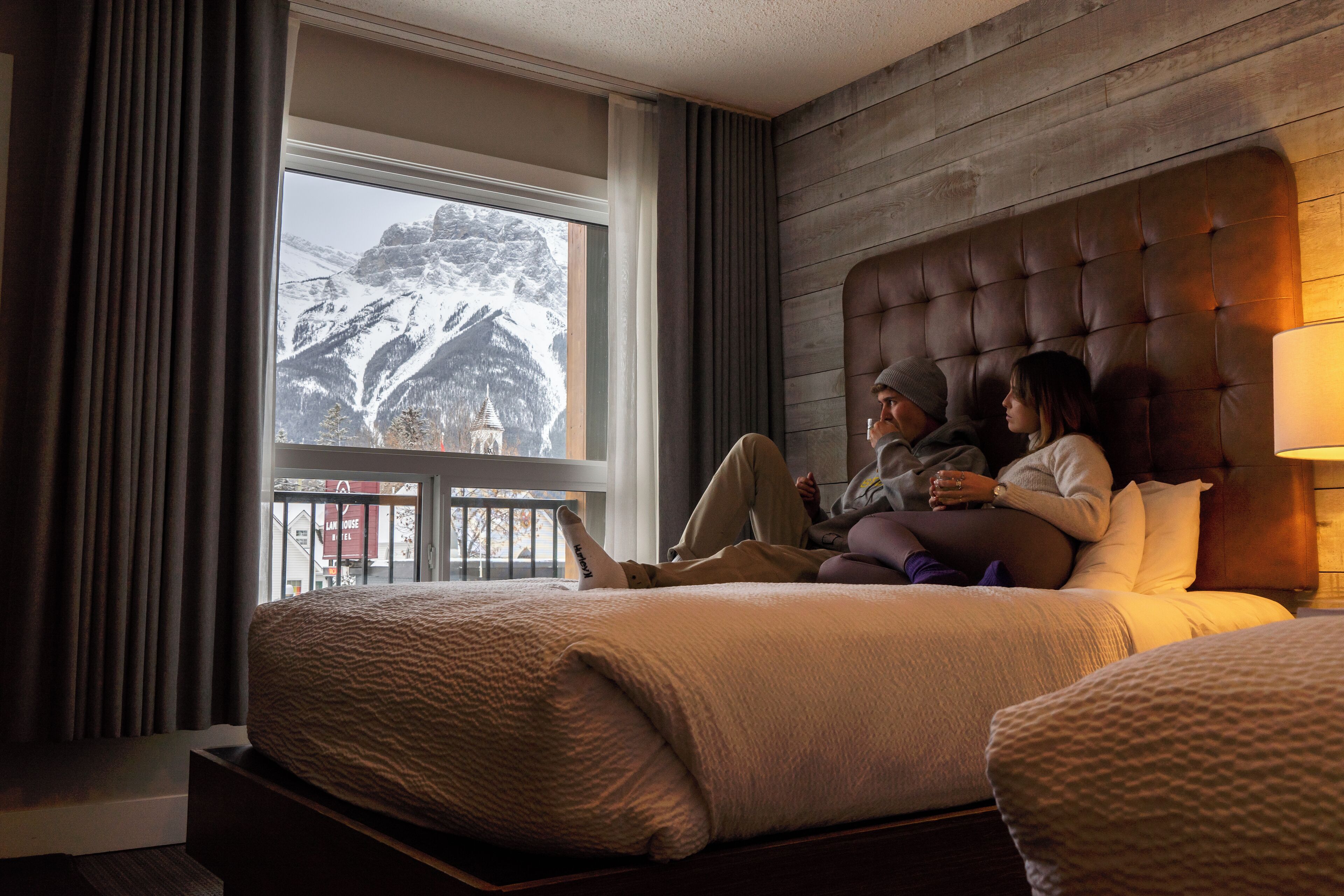 Habitación de hotel con un ventanal grande que ofrece vistas a la montaña, un cabecero capitonado y una lámpara de noche.