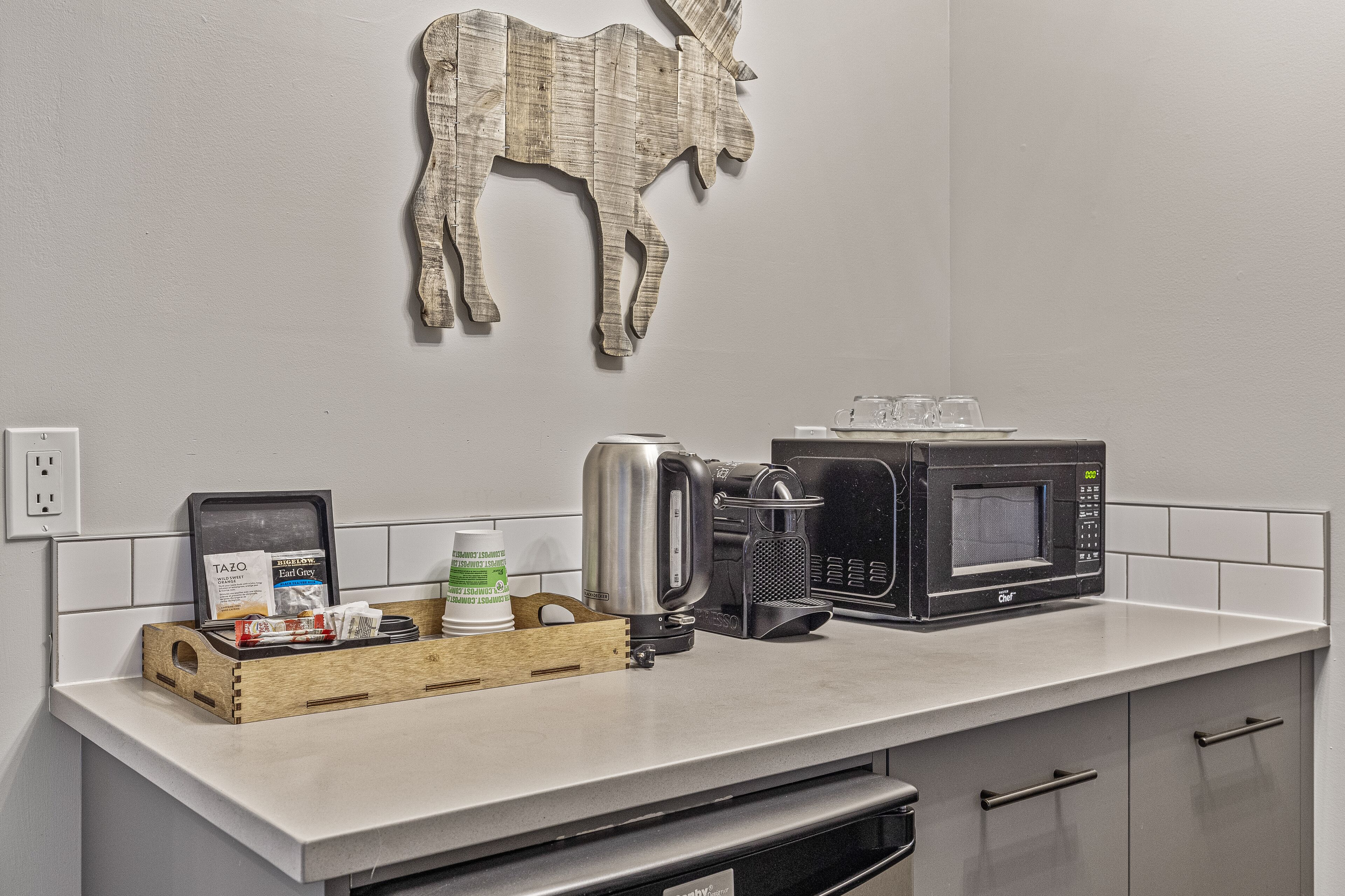 Un comptoir de cuisine avec une machine à café, un four à micro-ondes et un plateau en bois contenant des dosettes de café et une boîte de crème liquide.