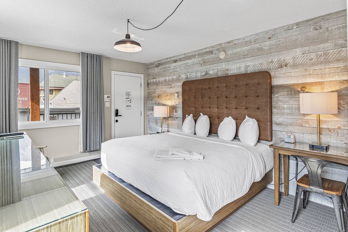 A hotel room with a large bed, a wooden headboard, two bedside lamps, a desk with a chair, and a view of the outside through a window.