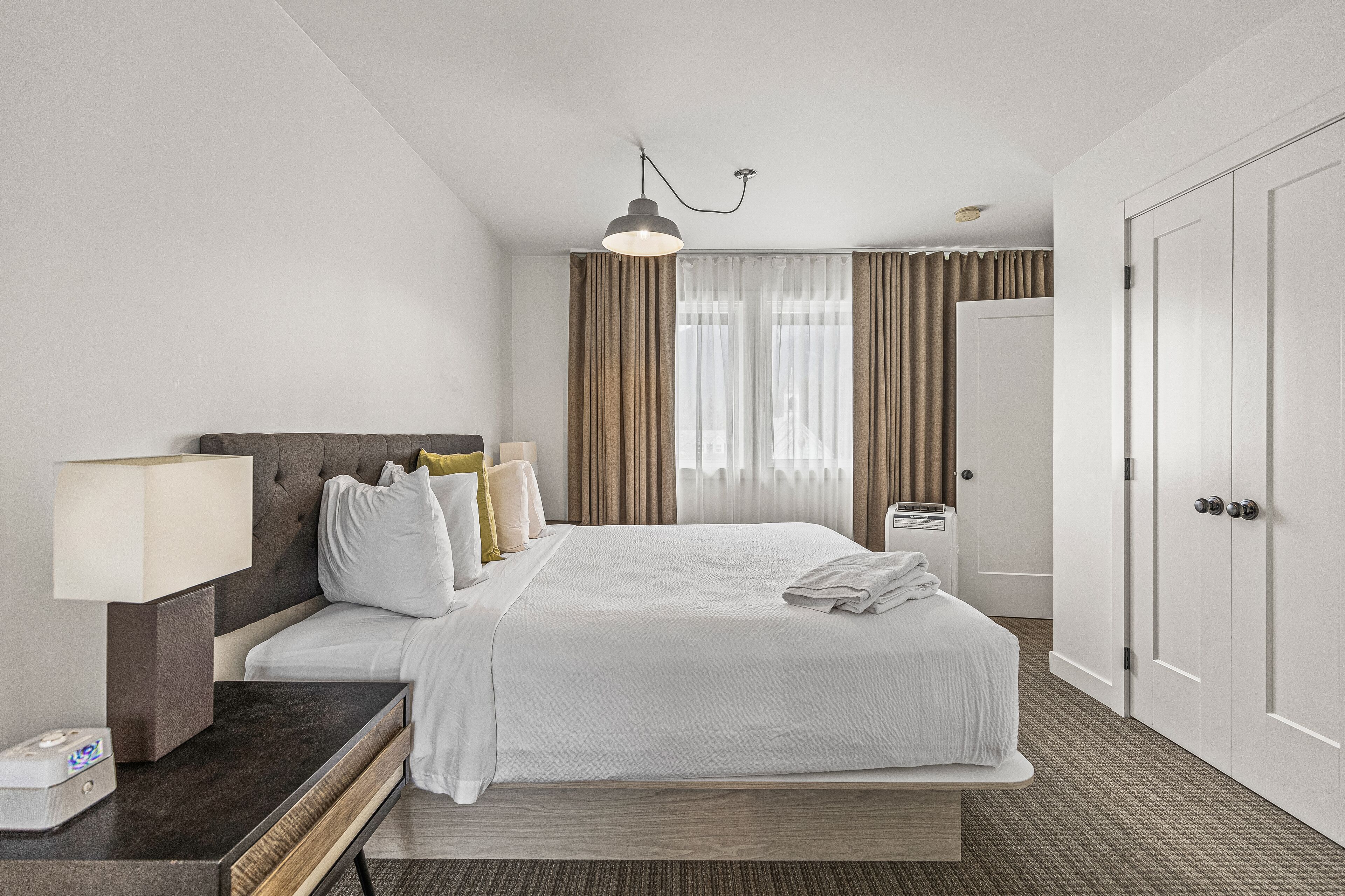 Una habitación de hotel con una cama grande, una mesita de noche con lámpara, un armario y una ventana con cortinas.