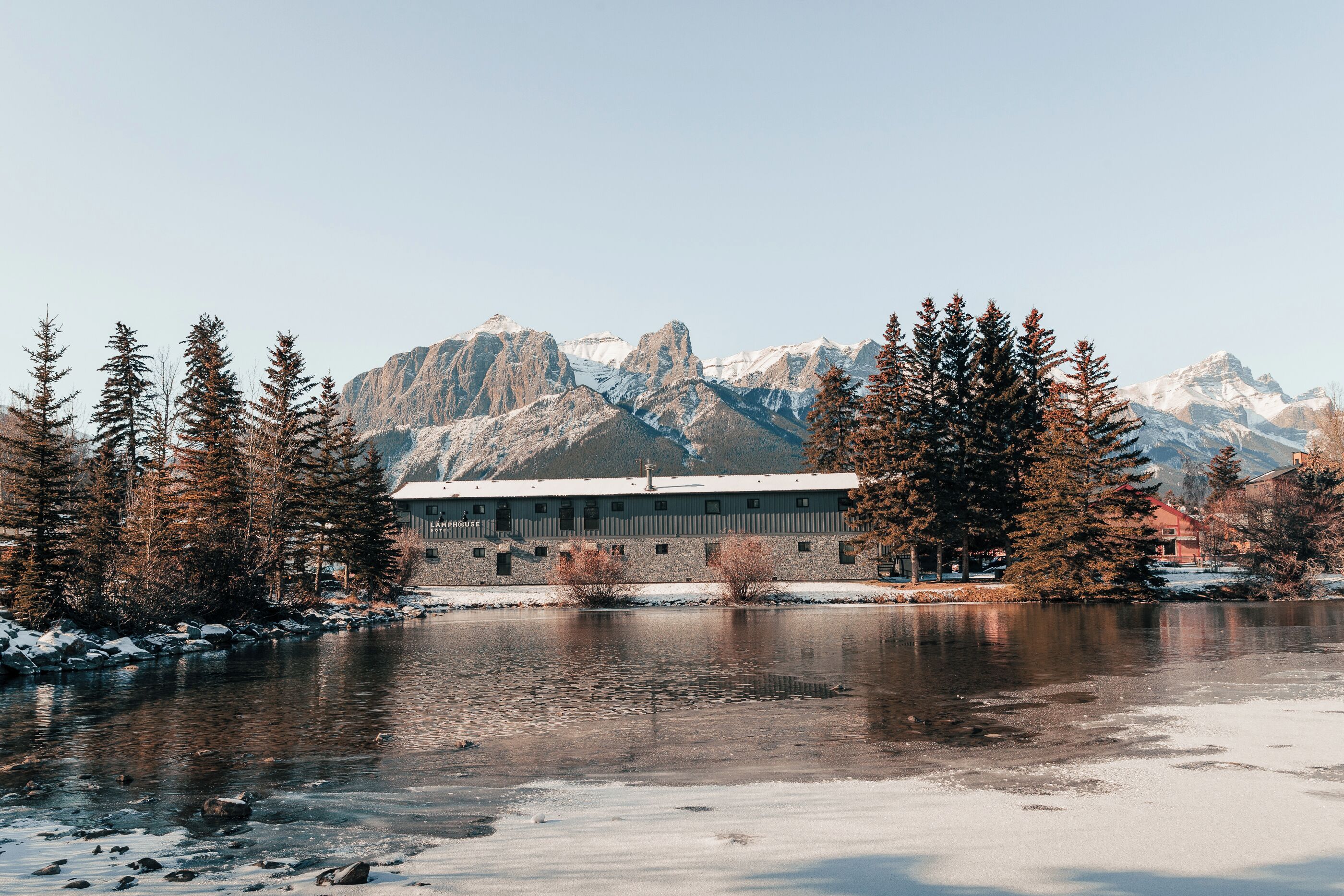 A building with a flat roof and a long, narrow structure, surrounded by trees and a frozen lake with mountains in the background.