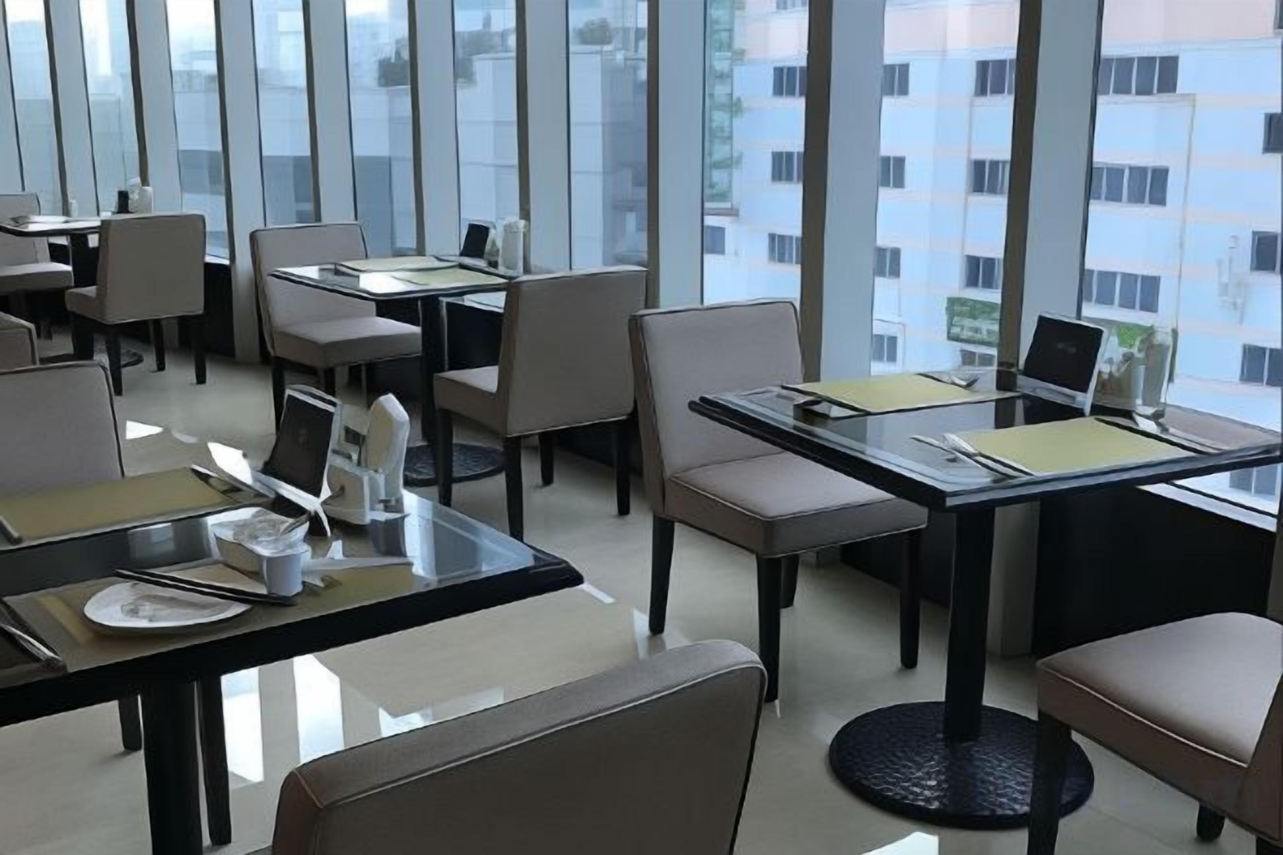 A modern dining area with tables and chairs, some with laptops, and large windows offering a view of the city.
