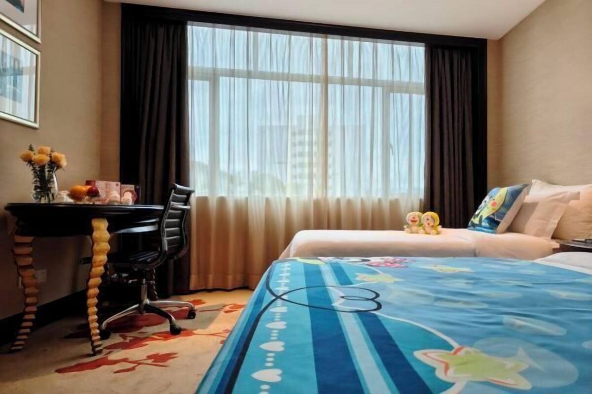 A hotel room with a bed, a desk with a chair, a vase with flowers, and a window with curtains.