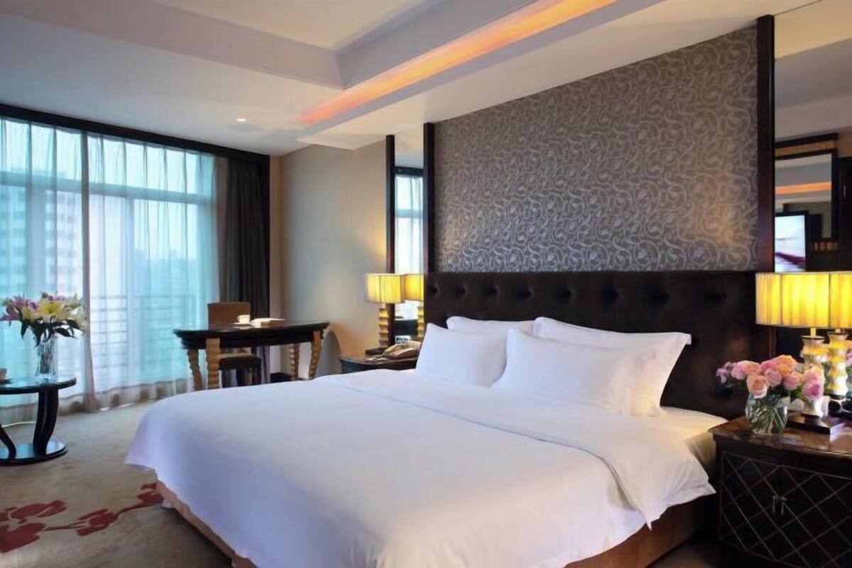 A hotel room with a large bed, a headboard with a patterned design, a desk with a lamp, and a view of the city through large windows.