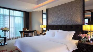 Habitación de hotel con una cama grande, cabecera con diseño estampado, escritorio con lámpara y vistas de la ciudad a través de amplios ventanales.