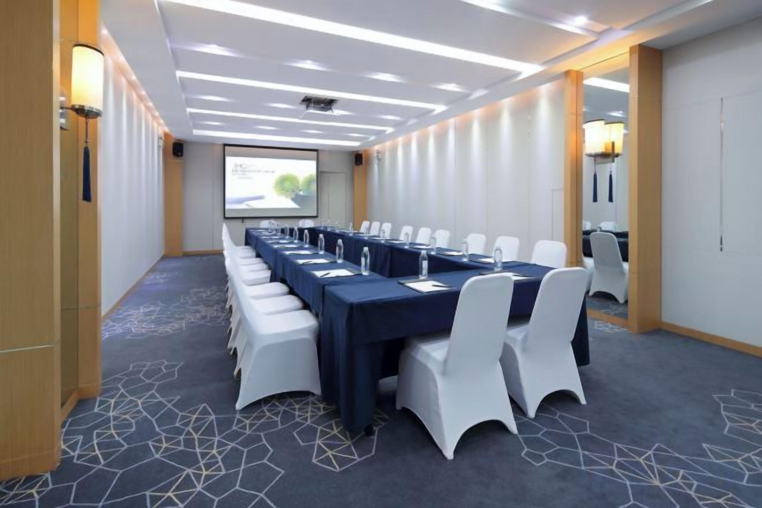 A conference room with a long table, white chairs, and a projector screen.