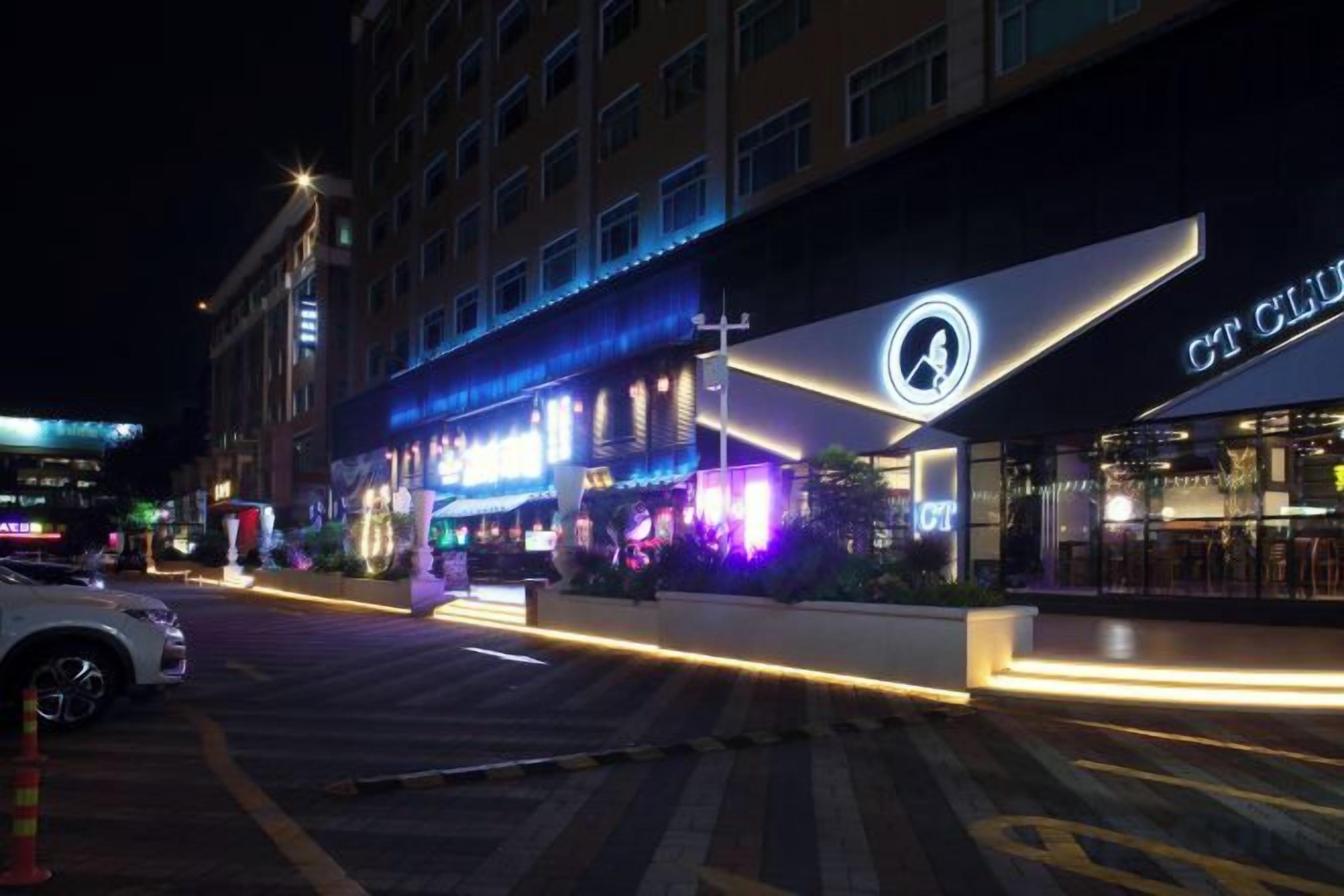 Entrada de hotel con diseño moderno, señalización iluminada y un área de estacionamiento visible.