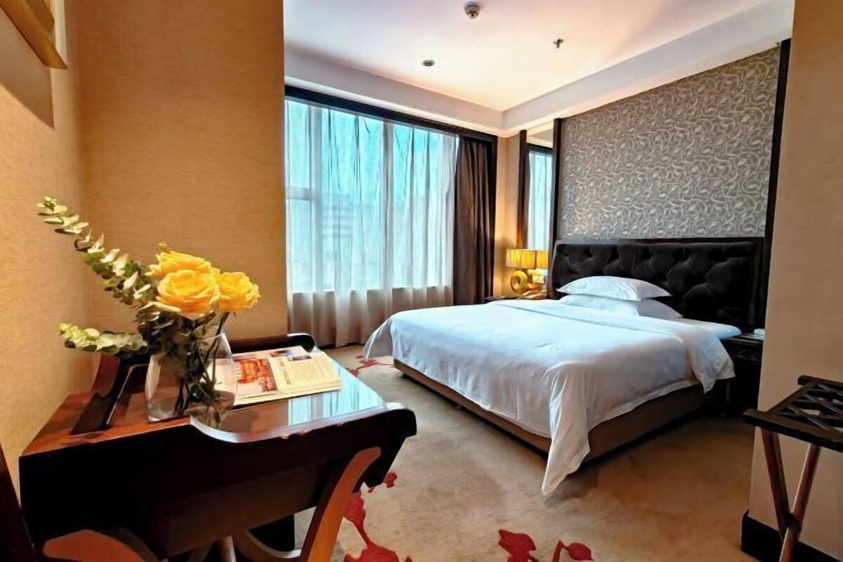 A hotel room with a bed, a desk with a chair, a vase with flowers, and a window with curtains.