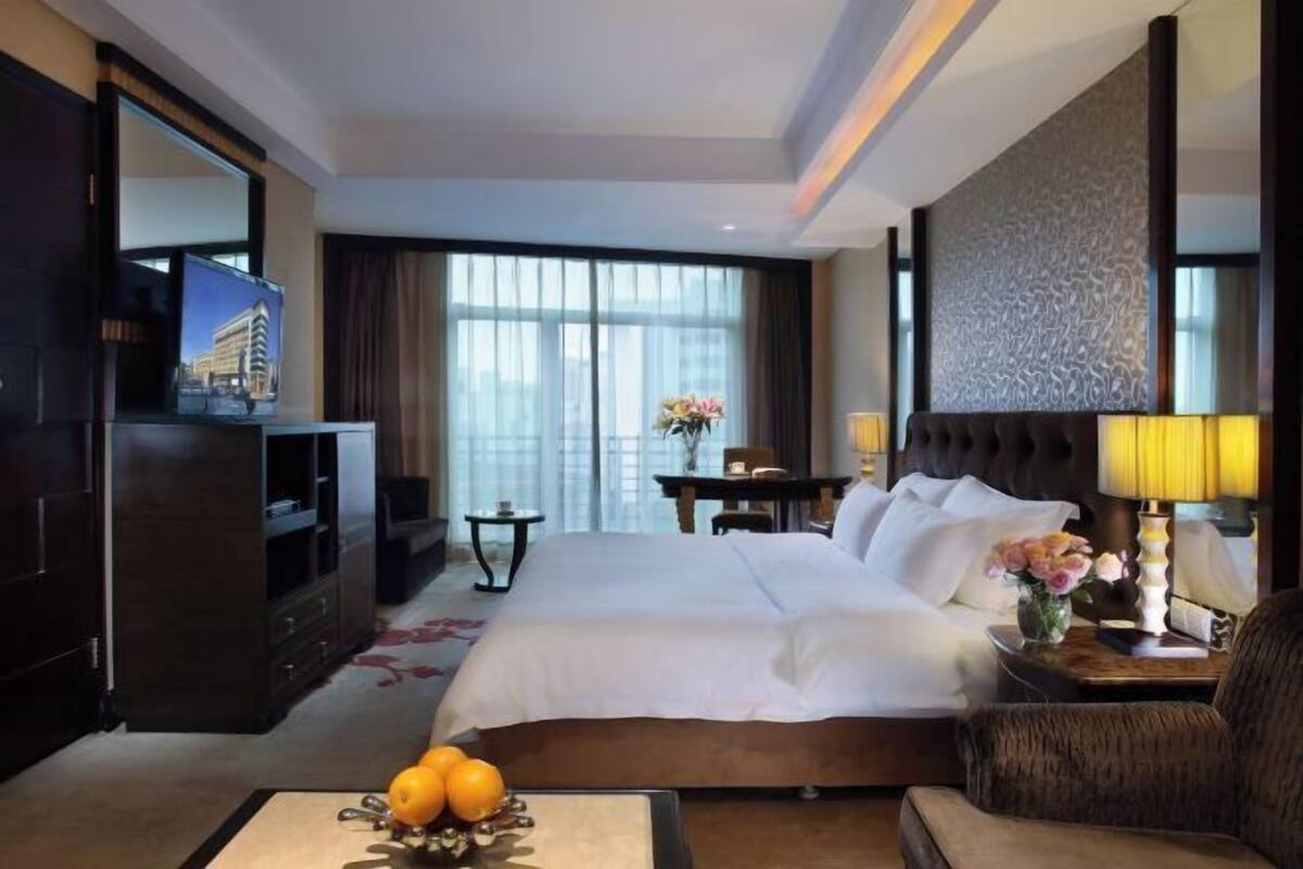 A hotel room with a large bed, a desk, a television, and a view of the city through large windows.