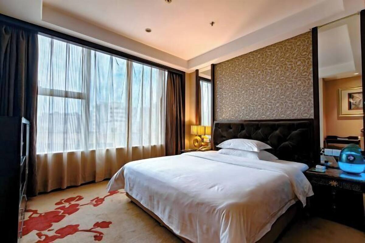 A hotel room with a large bed, a nightstand, a lamp, and a view of the outdoors through large windows.