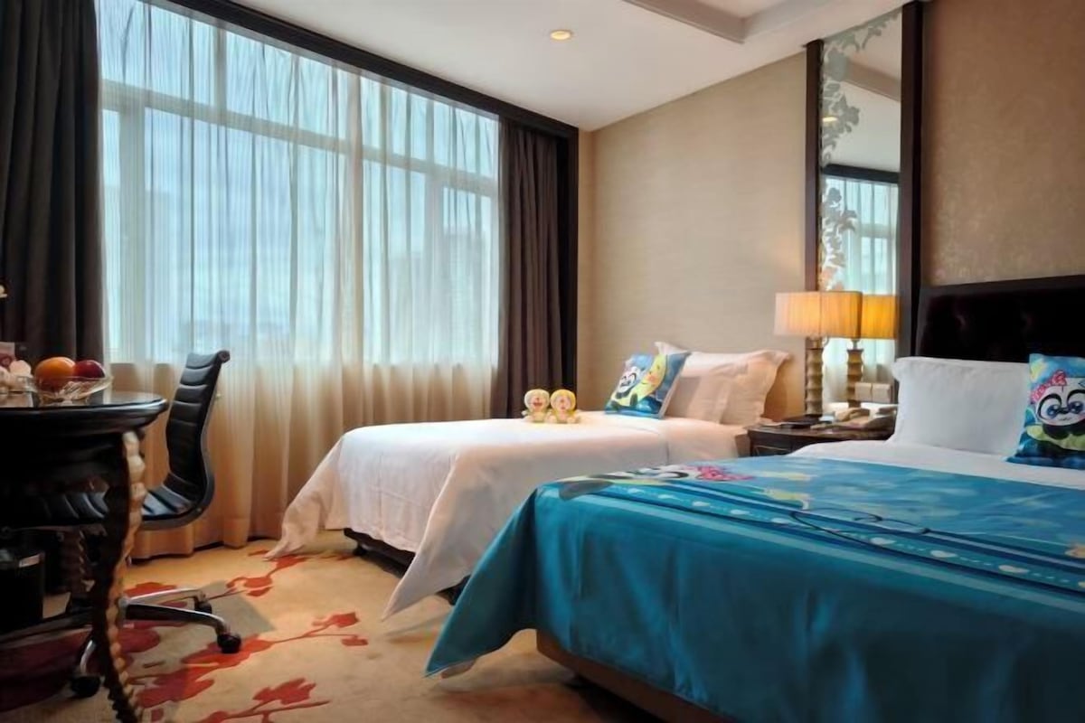 A hotel room with two beds, a desk with a chair, a large window with curtains, and a mirror on the wall.