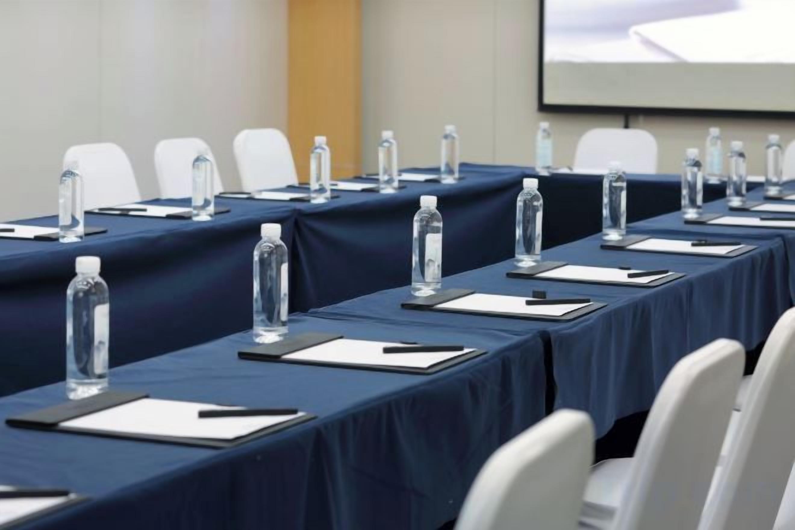 Un salón de conferencias con mesas dispuestas para una reunión, cada una con una botella de agua, un bloc de notas y un bolígrafo.