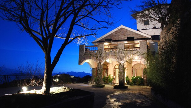 A stone building with arched doorways and a balcony, surrounded by trees and a mountain backdrop.