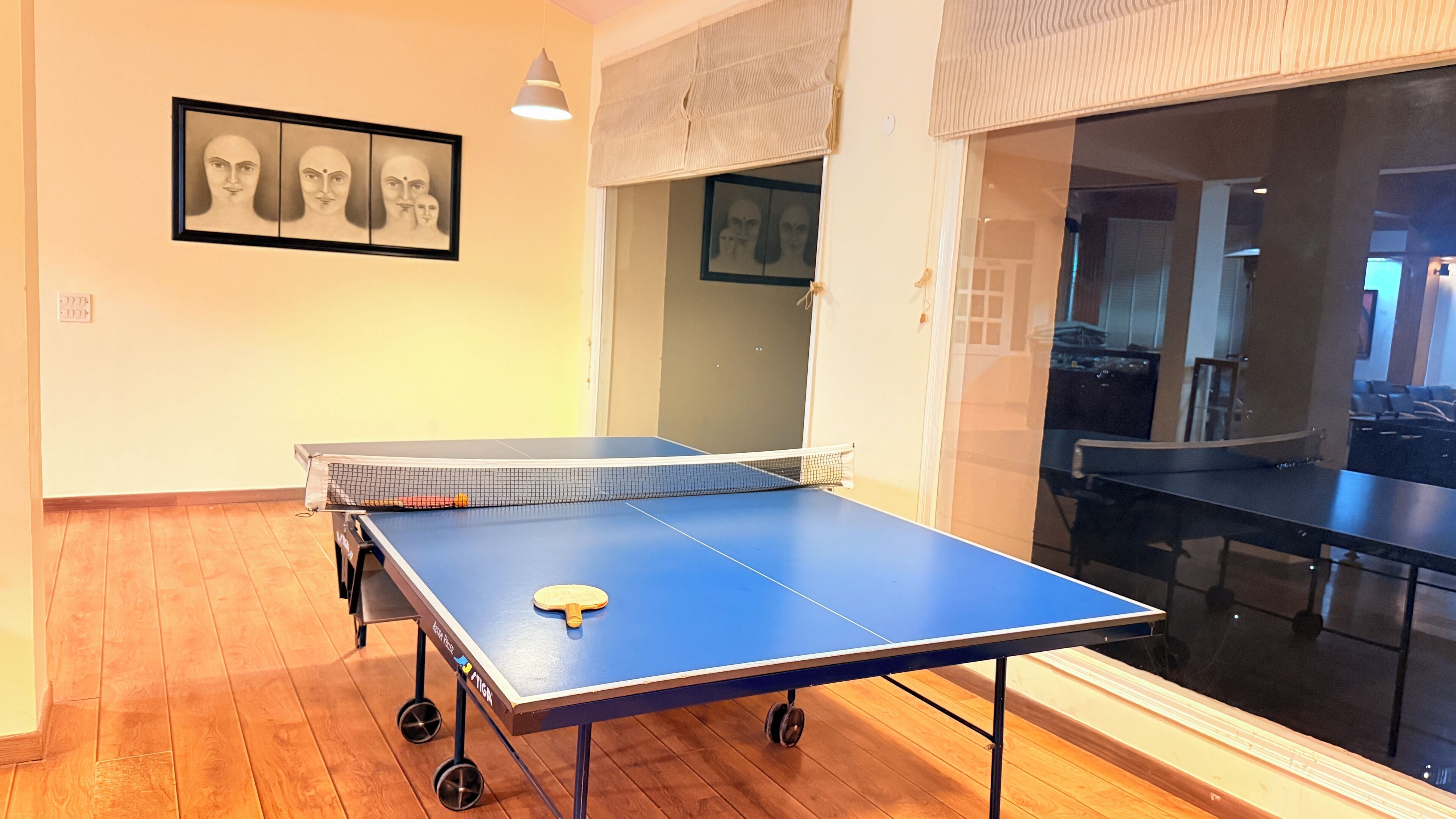A ping pong table in a room with wooden flooring, a framed artwork on the wall, and a window with blinds.