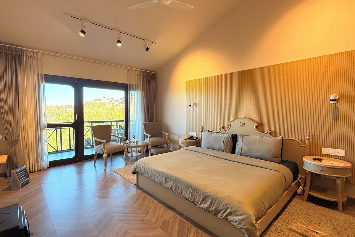 A spacious hotel room with a large bed, a desk, and a chair. There is a balcony with a view of the outdoors.
