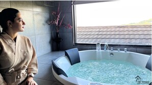 A person sitting by a large indoor hot tub with a view of a tiled roof outside.
