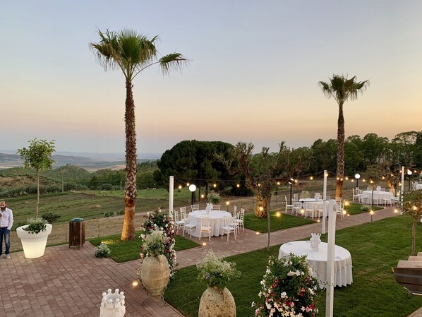 An outdoor wedding venue with round tables and chairs, surrounded by palm trees and potted plants.