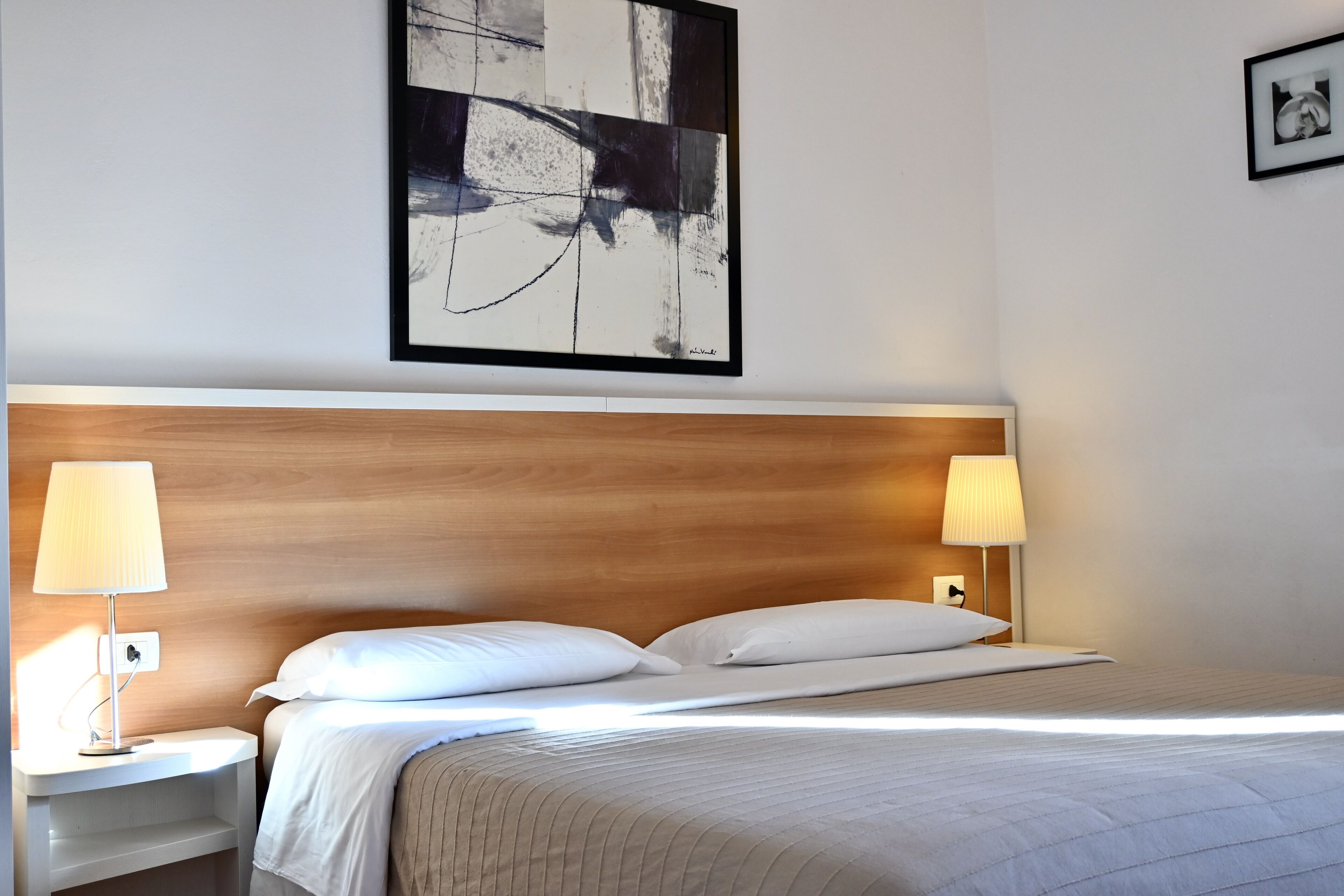 Una cama bien hecha con ropa blanca, dos lámparas de noche y un cuadro abstracto grande en la pared.