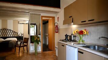 A compact kitchen with a refrigerator, stove, sink, and a vase of flowers. A wooden chair and a bed are visible in the adjacent room.