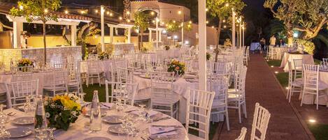 An outdoor dining area with round tables set for a meal, surrounded by white chairs and decorated with flowers.