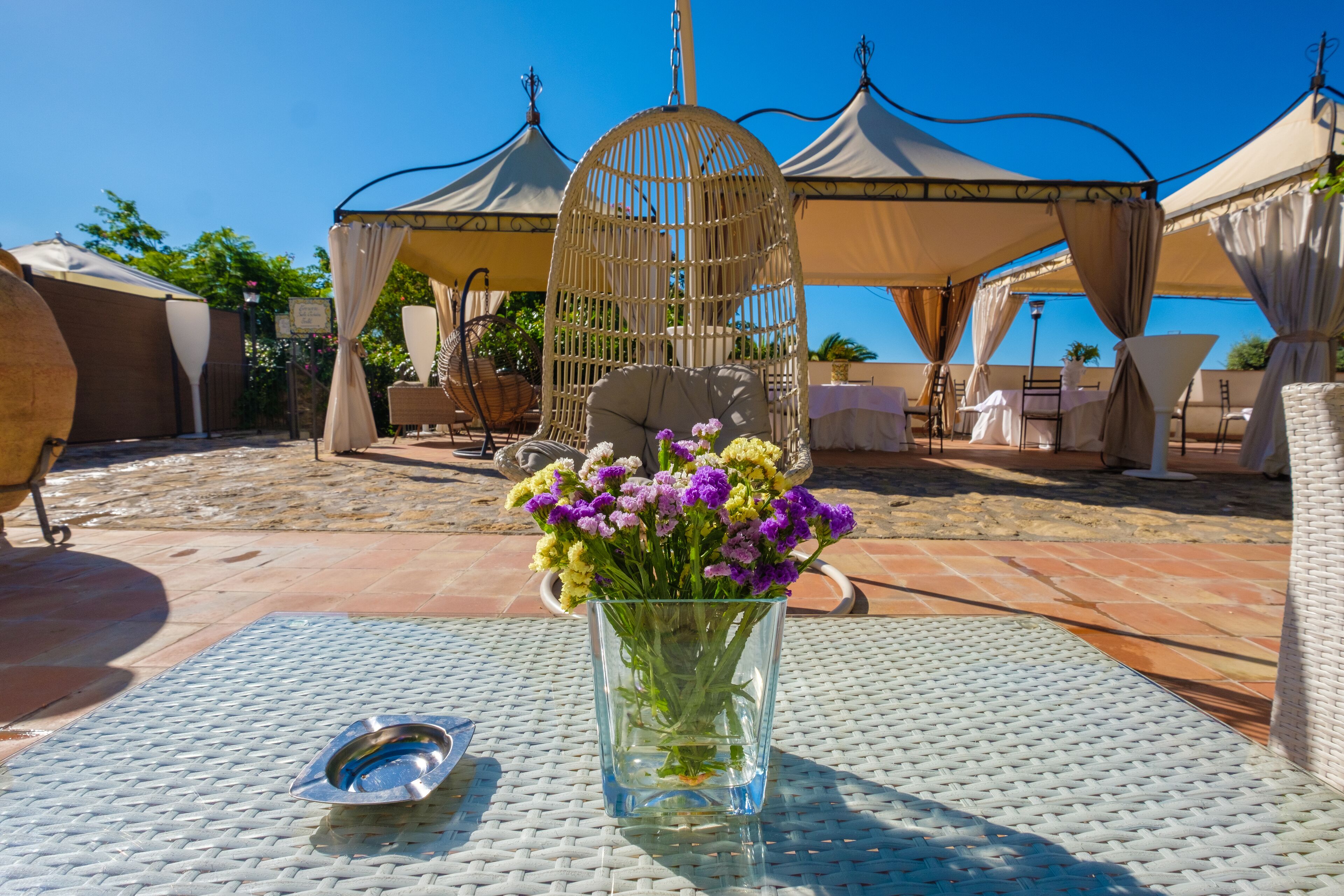 Eine Glasvase mit Blumen auf einem Tisch, ein Vogelkäfig und eine überdachte Sitzgelegenheit mit Stühlen und Tischen.