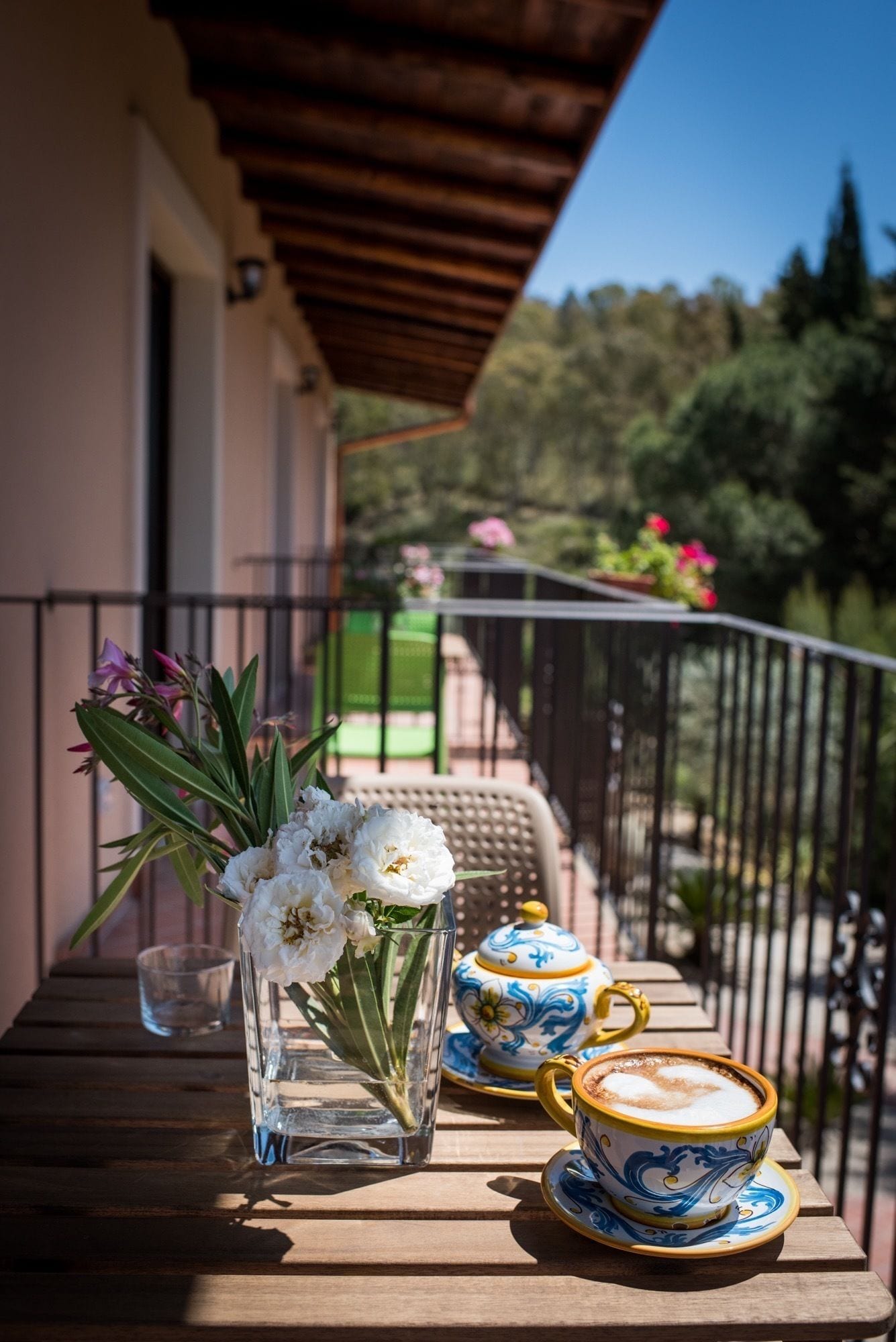 Un balcón con una mesa de madera, un florero de cristal transparente con flores blancas y dos tazas de cerámica con un diseño azul y amarillo.