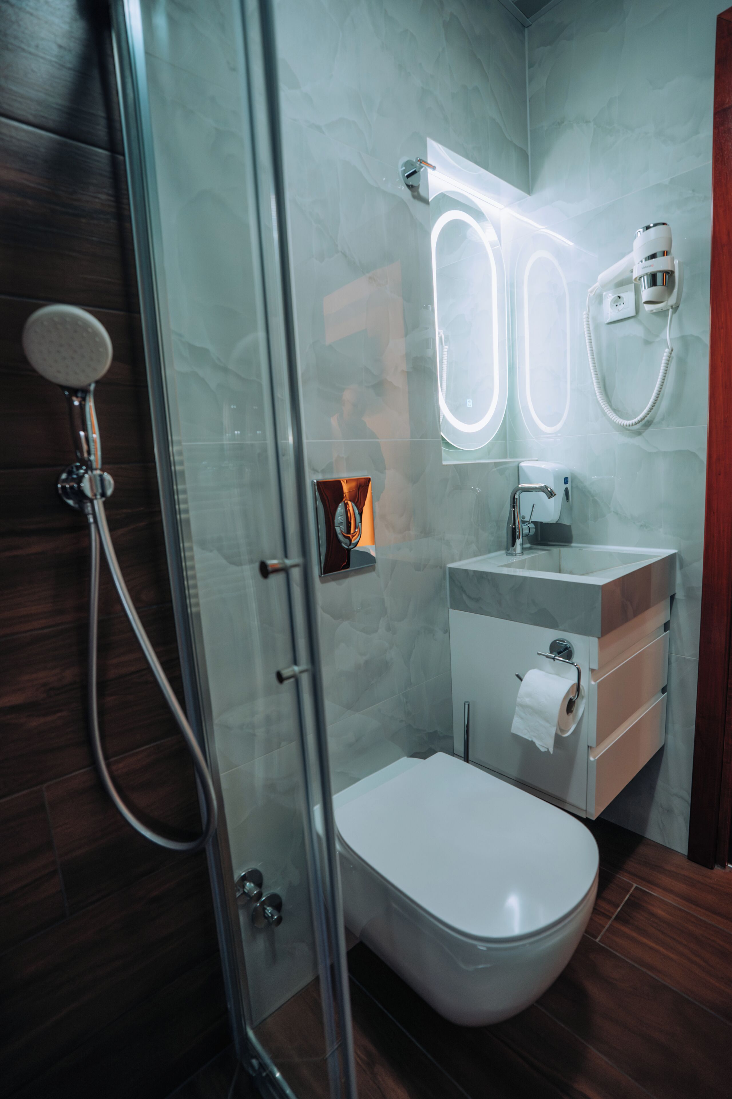 Une salle de bain moderne avec une douche à parois de verre, des toilettes blanches, un lavabo en marbre et un distributeur de serviettes.