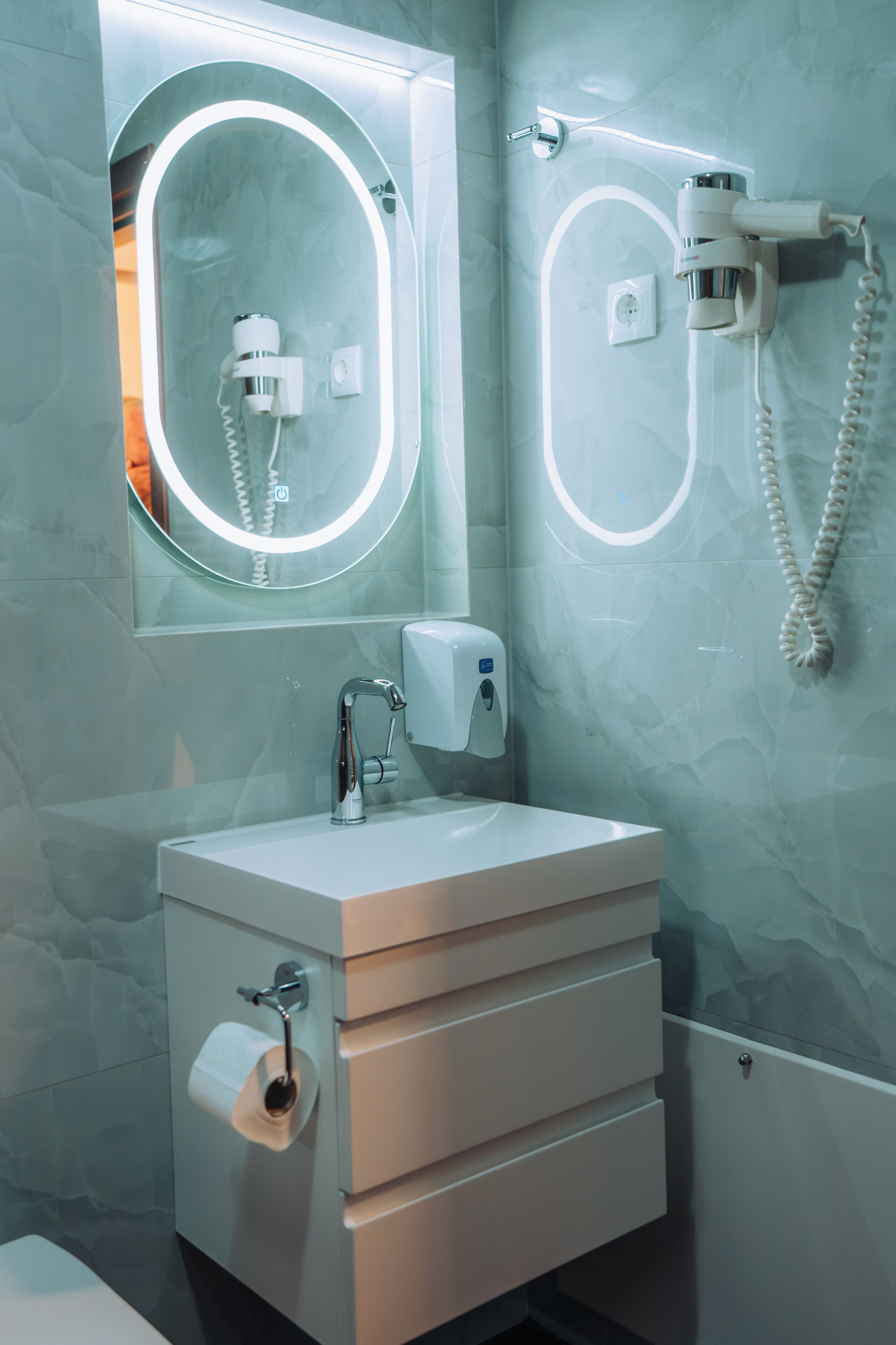 A bathroom with a mirror, a sink, a towel dispenser, a hairdryer, and a shower head.