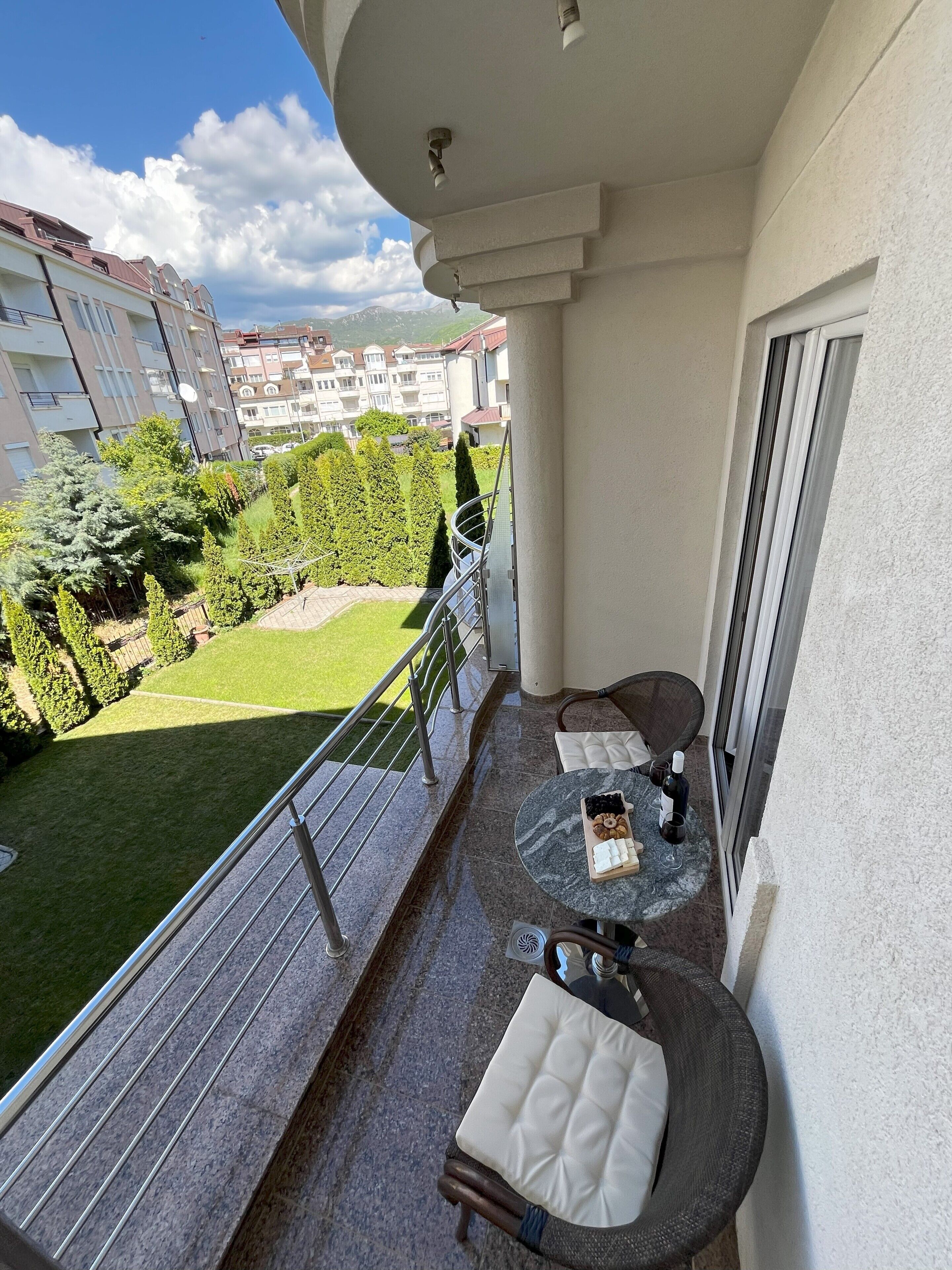 A balcony with a marble table, two cushioned chairs, a small garden view, and a building in the background.