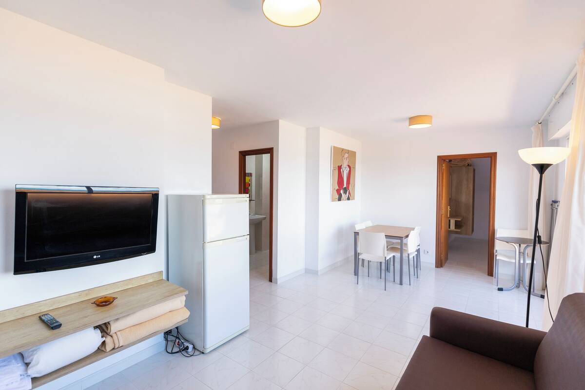 A modern living room with a flat-screen TV, a white refrigerator, a wooden console table, a brown sofa, and a dining area with a table and chairs.