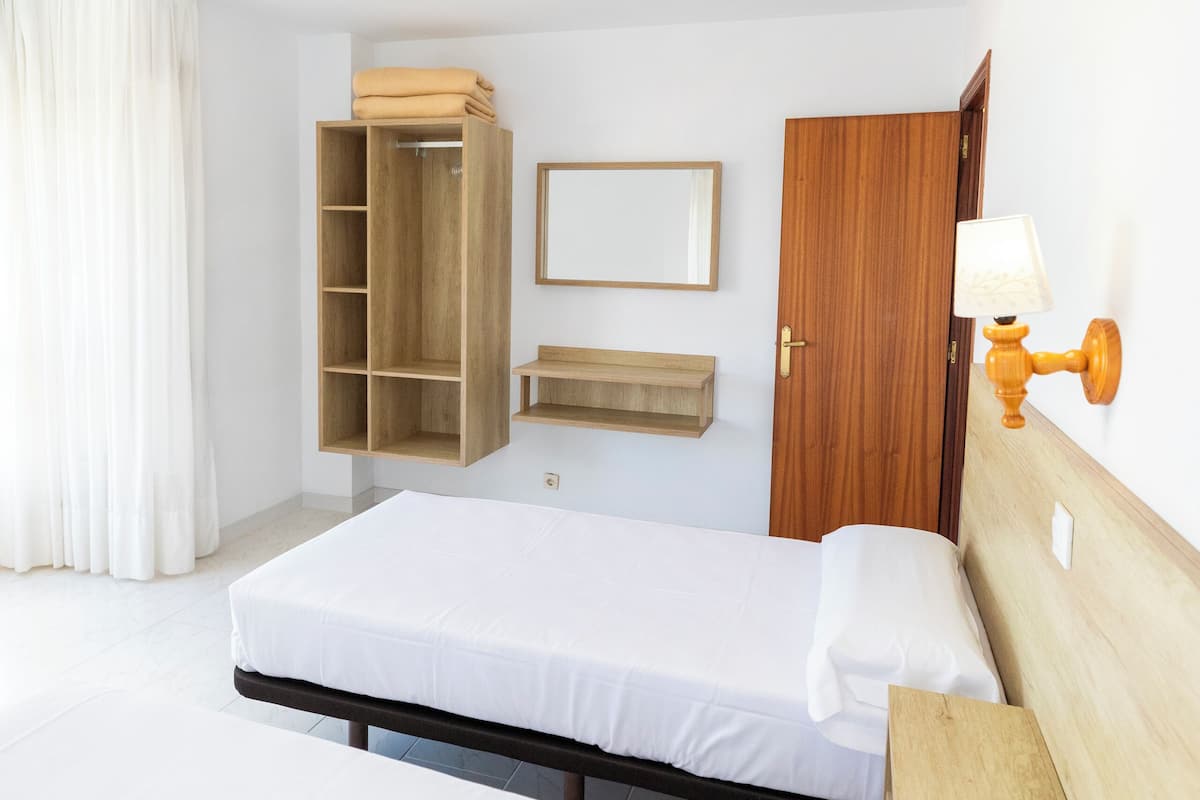 A hotel room with a bed, a wooden wardrobe, a bedside table, a mirror, and a wall-mounted lamp.
