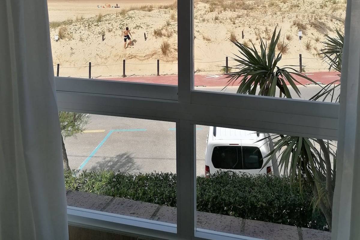 A beach view through a window, including a sandy area, a white van, and a building in the distance.
