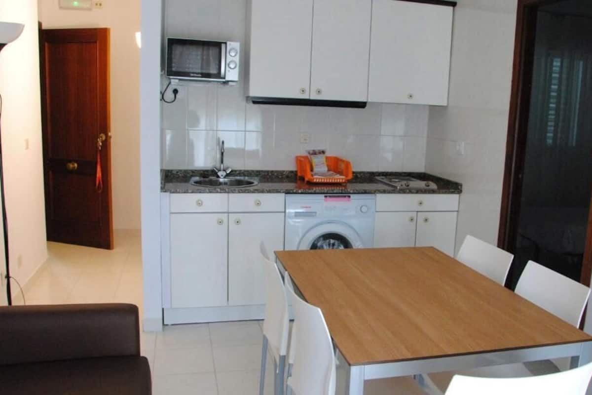 A compact kitchen with a microwave, sink, and washing machine, a dining table with chairs, and a wooden door.
