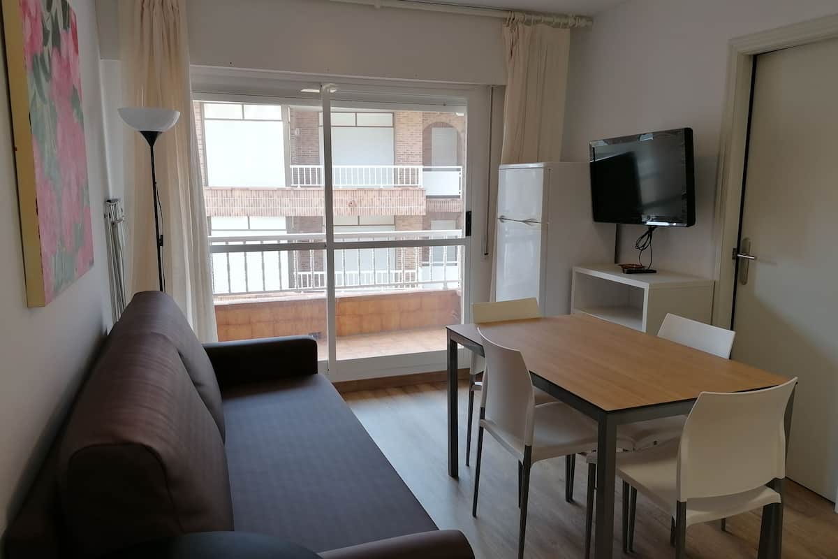 A modern living room with a sofa, a dining table with chairs, a TV mounted on the wall, and a balcony with a view of buildings.