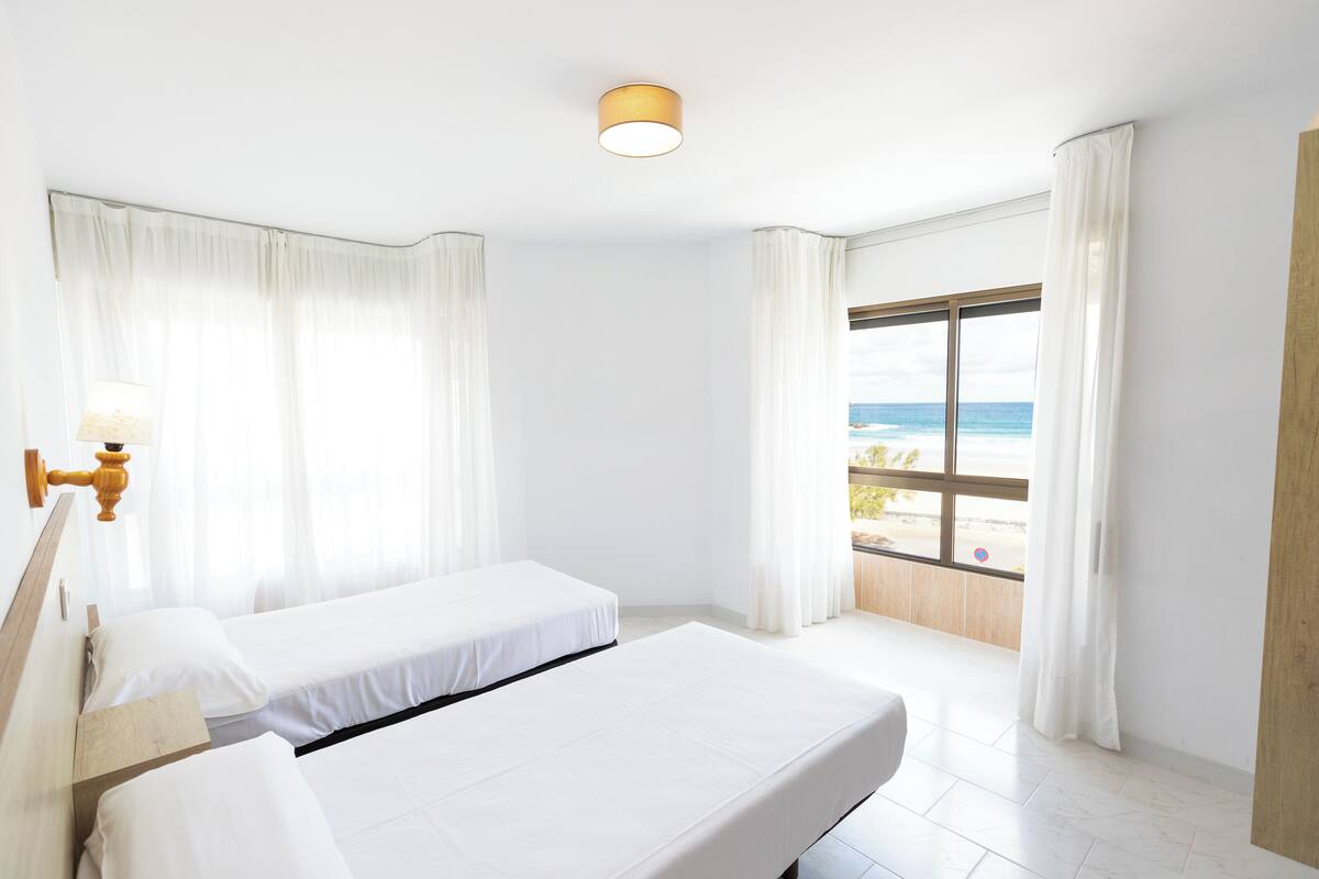 A hotel room with two beds, a large window with a view of the beach, and a ceiling light.
