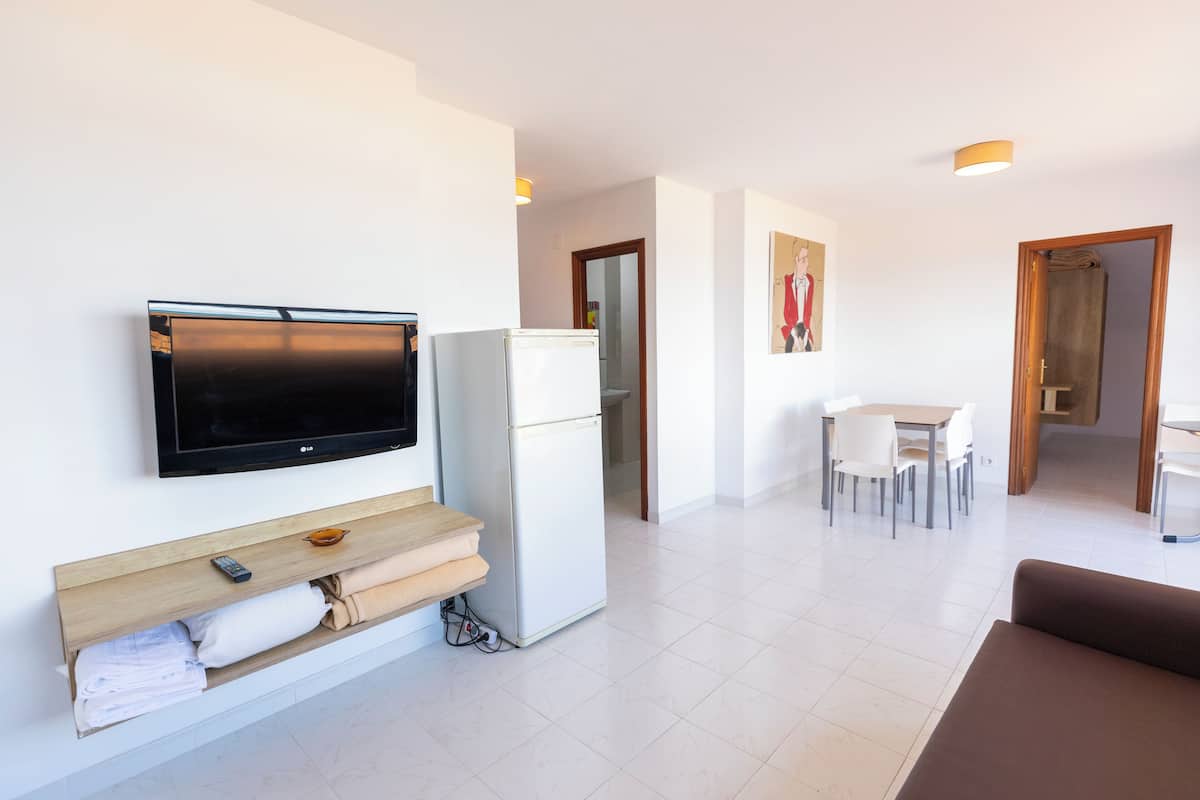 A modern living room with a flat-screen TV, a white refrigerator, a wooden shelf with towels, a brown sofa, a dining area with a table and chairs, and a hallway leading to a door.
