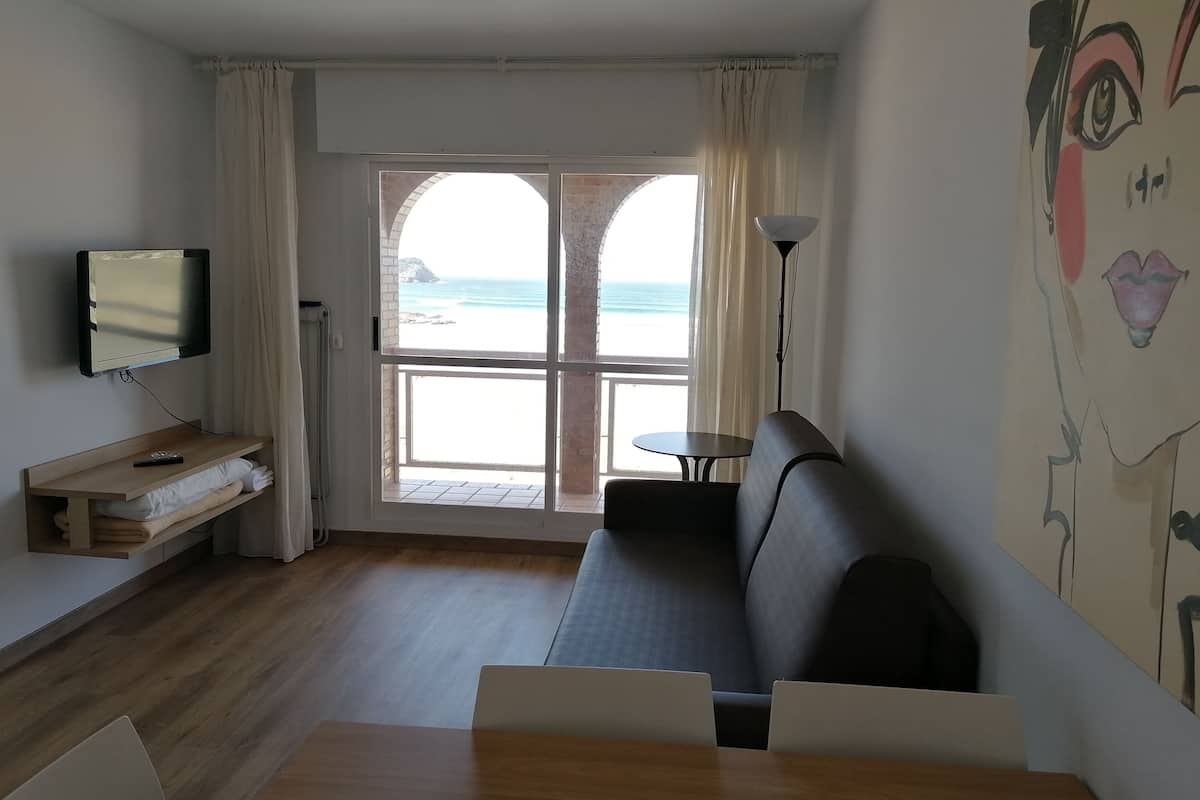 A room with a wooden floor, a sofa, a table, a TV on a stand, a window with curtains, and a painting on the wall.