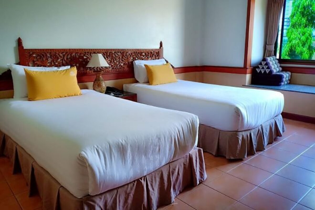 Une chambre d’hôtel avec deux lits, chacun agrémenté de draps blancs et d’oreillers jaunes. On y trouve une tête de lit en bois ornée de motifs complexes, une table de chevet avec une lampe et une fenêtre donnant sur un paysage verdoyant.