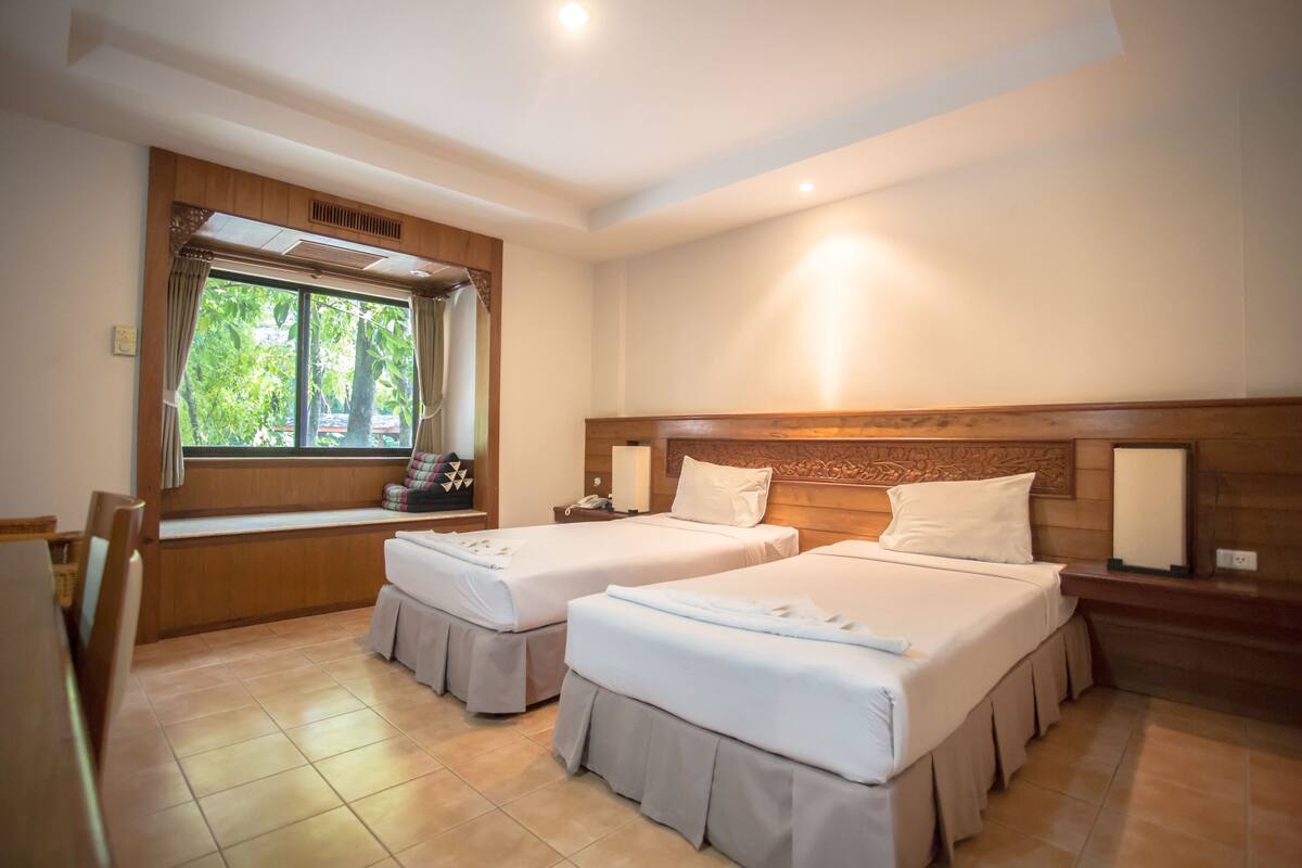 Ein Hotelzimmer mit zwei Betten, einem Fenster mit Vorhängen, einem hölzernen Kopfteil und einem Nachttisch mit Lampe.