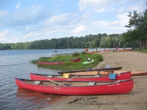 A lakeside setting with canoes lined up on the shore, a sandy beach, and a forested background.