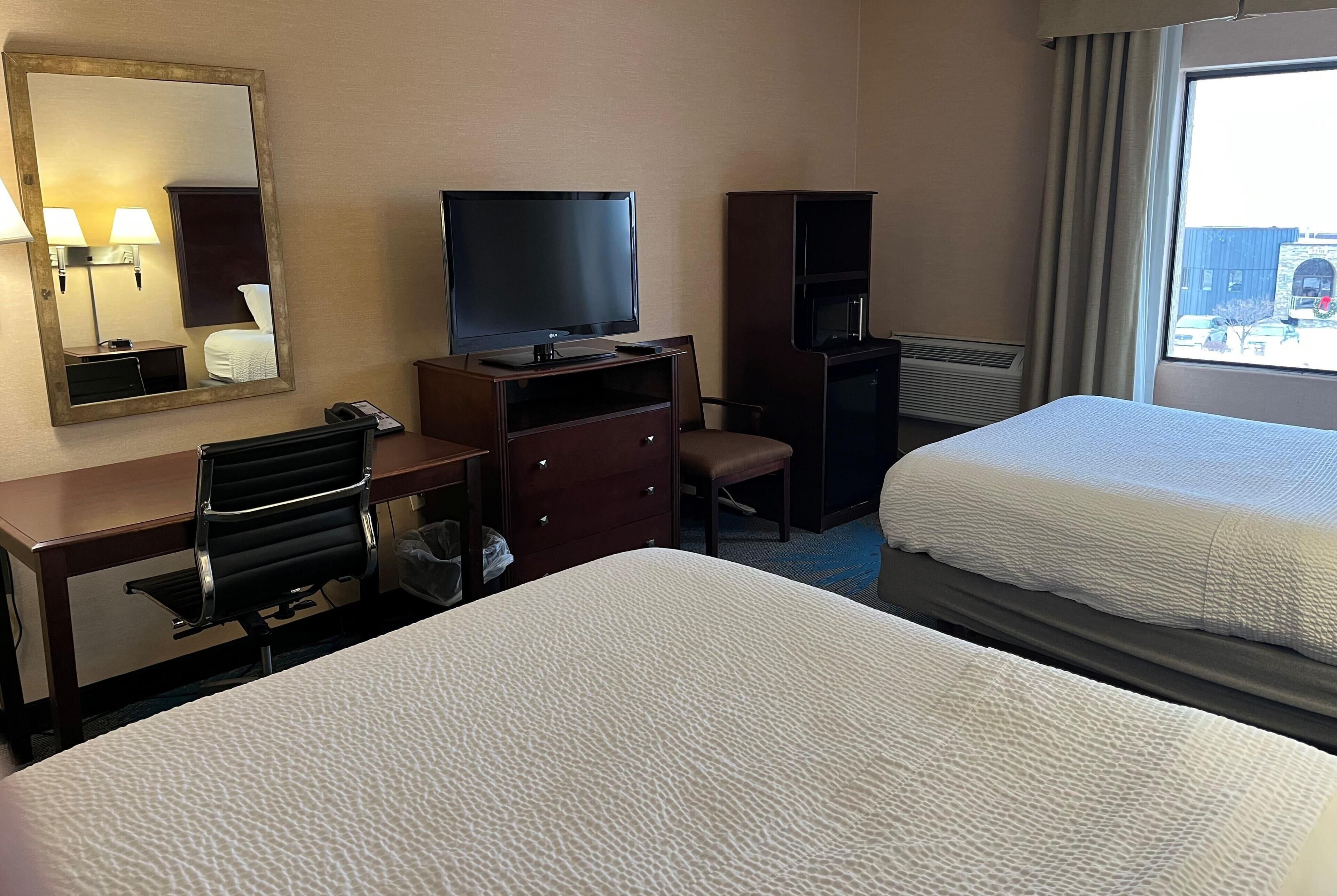 A hotel room with two beds, a desk with a chair, a television, a mirror, and a window with curtains.