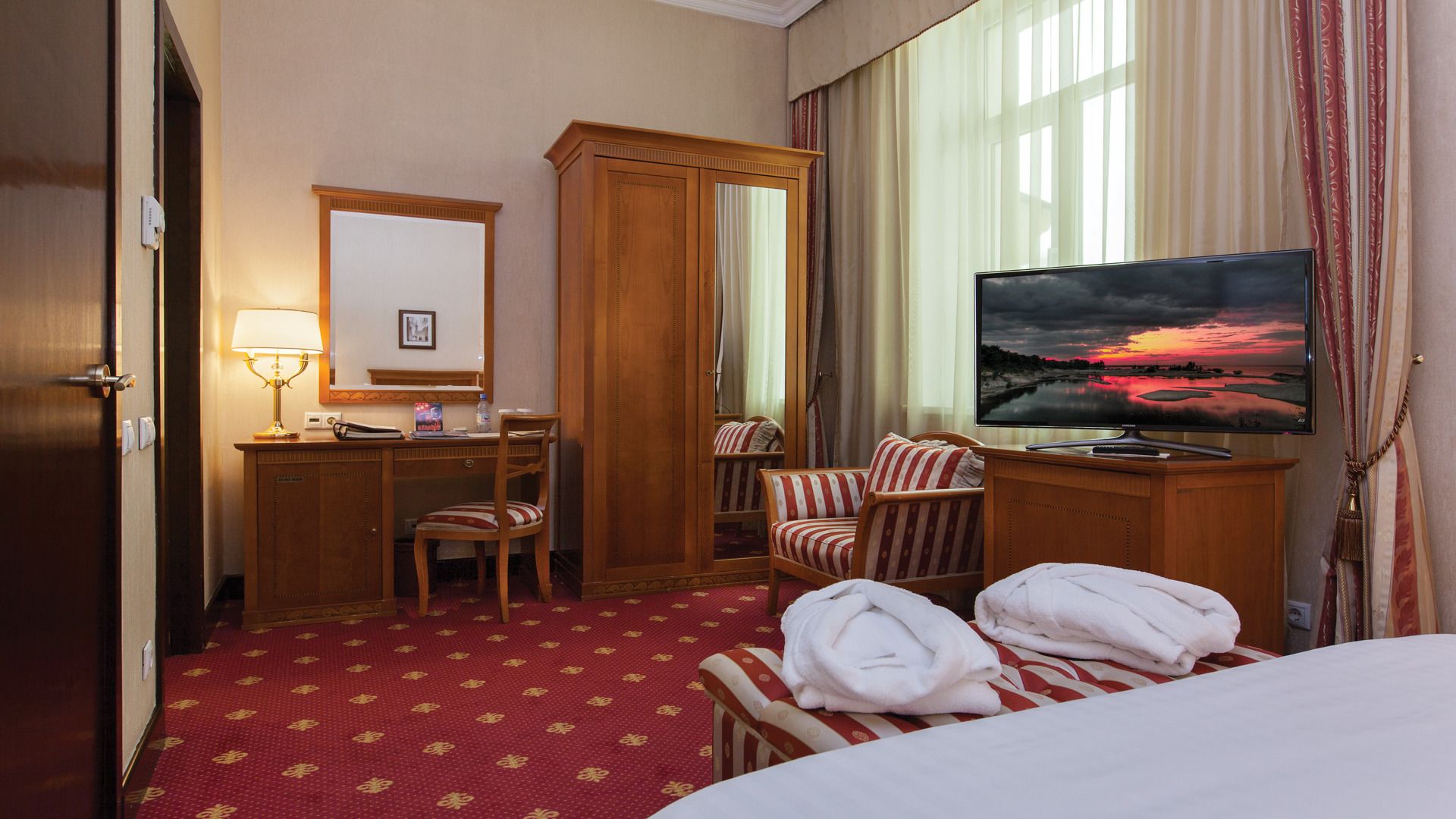 A hotel room with a bed, a desk with a lamp, a TV, a chair, and a wardrobe.