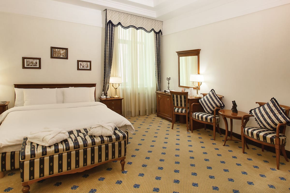A hotel room with a large bed, two bedside tables with lamps, a wooden dresser, two upholstered chairs, a mirror, and three framed pictures on the wall.