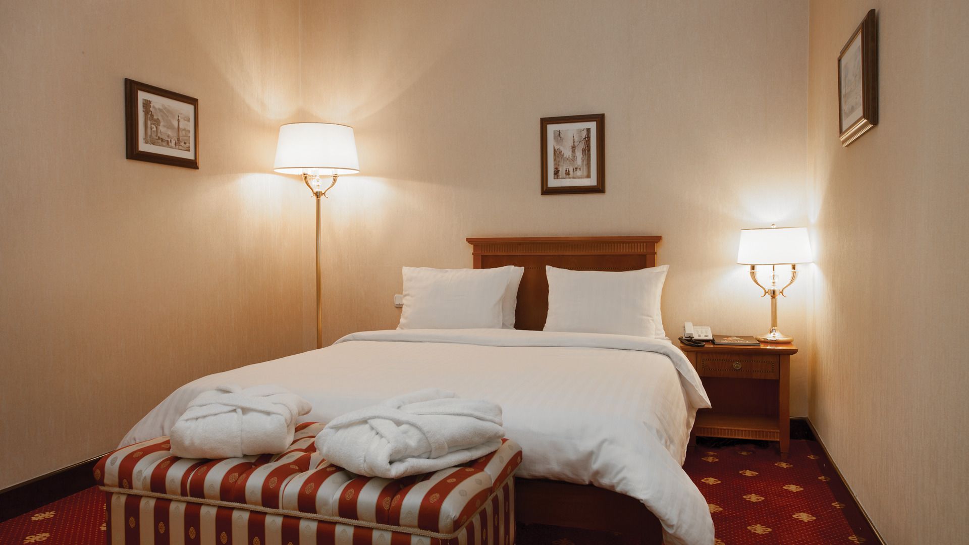 Habitación de hotel con una cama bien hecha, dos lámparas de noche, una mesita con un teléfono y un cuadro enmarcado en la pared.